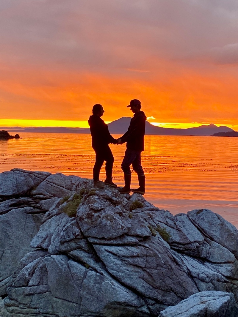 Couple stands on a rock at the Sitka Halibut Point Recreation Area with the sunset in the background. 